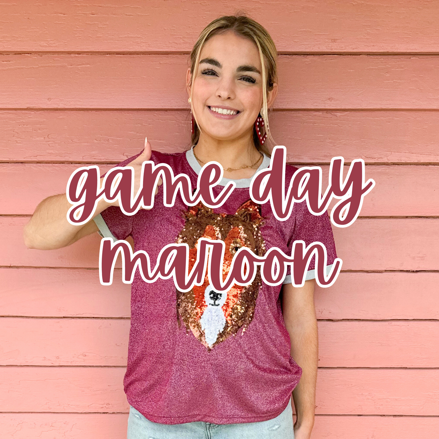 A smiling blonde woman poses against a pink wooden wall, wearing a maroon game day glitter tee featuring a sequin Reveille design. "Game Day Maroon" is overlaid in bold maroon script, highlighting a stylish, spirited top perfect for supporting your team.