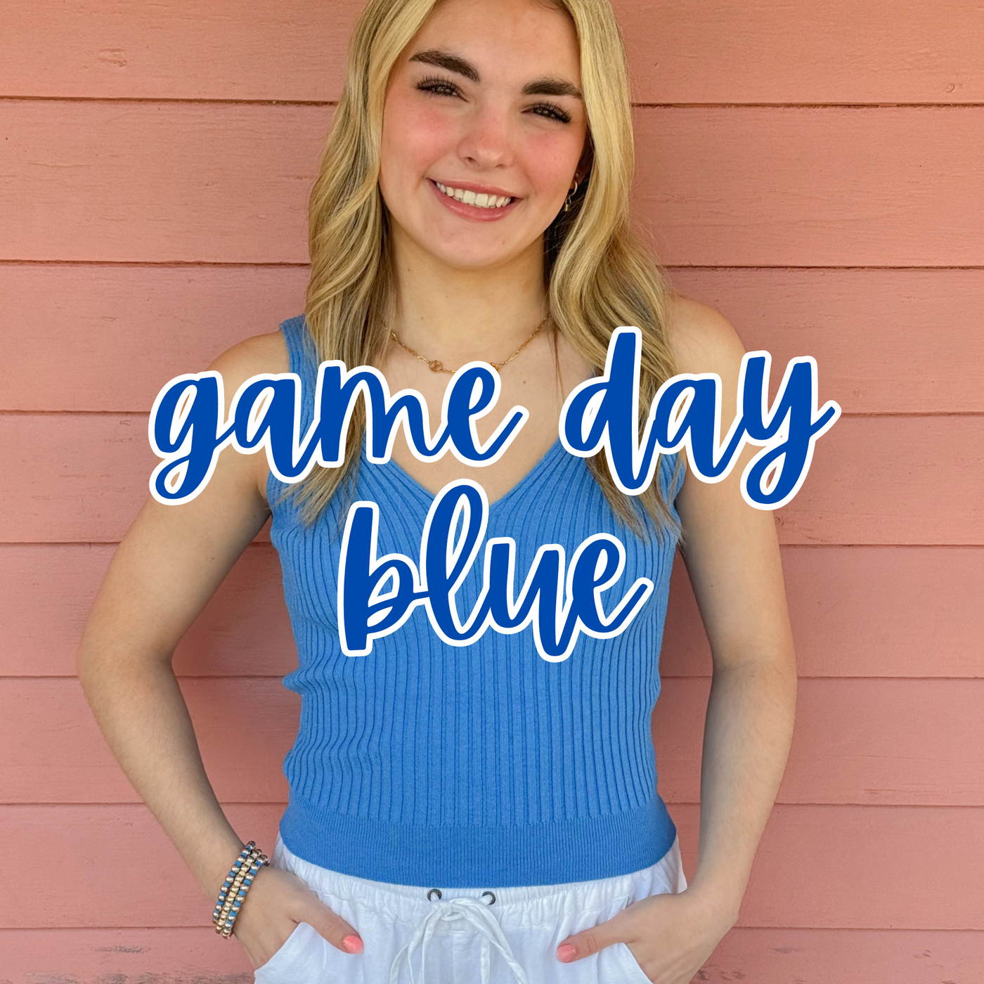 A smiling blonde woman poses against a pink wooden wall, wearing a ribbed blue V-neck tank top and white drawstring shorts. "Game Day Blue" is overlaid in bold blue script, highlighting a stylish and sporty outfit perfect for showing team spirit.