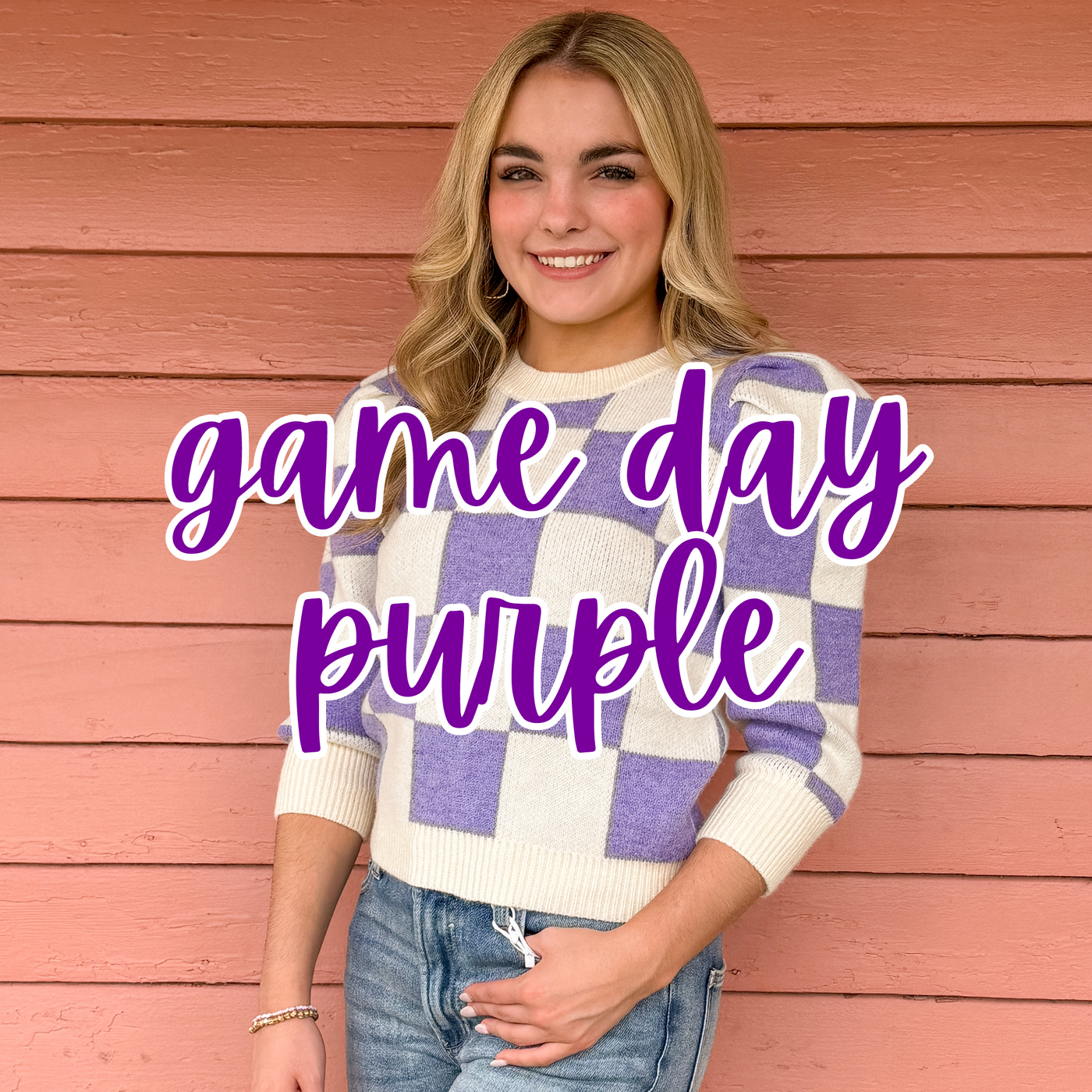 A smiling blonde woman poses against a pink wooden wall, wearing a purple and white checkered sweater with puff sleeves and a denim skirt. "Game Day Purple" is overlaid in bold purple script, showcasing a stylish outfit perfect for supporting your team.