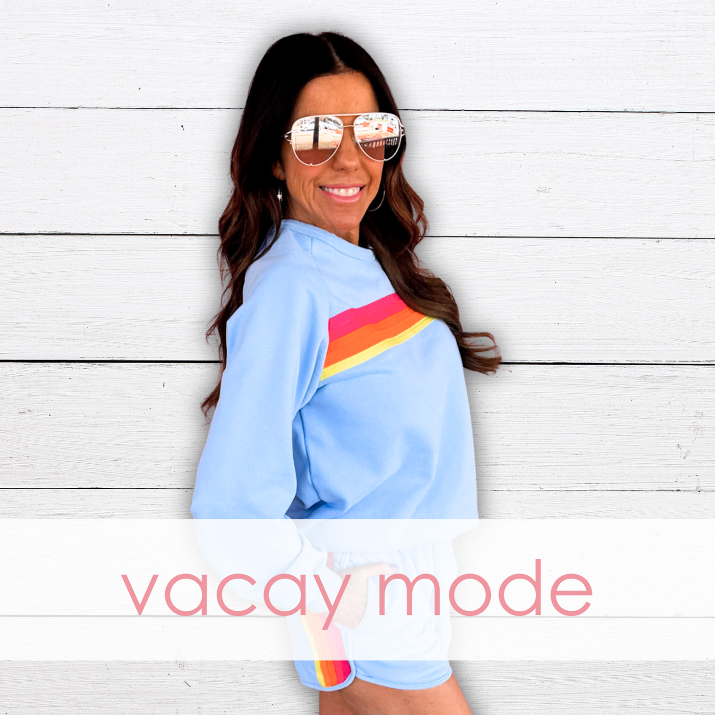 A smiling woman with wavy brown hair poses against a white shiplap wall, wearing a light blue sweatshirt, white shorts with rainbow stripes, and aviator sunglasses. "Vacay Mode" is overlaid in pink text, highlighting a fun, relaxed vacation look.