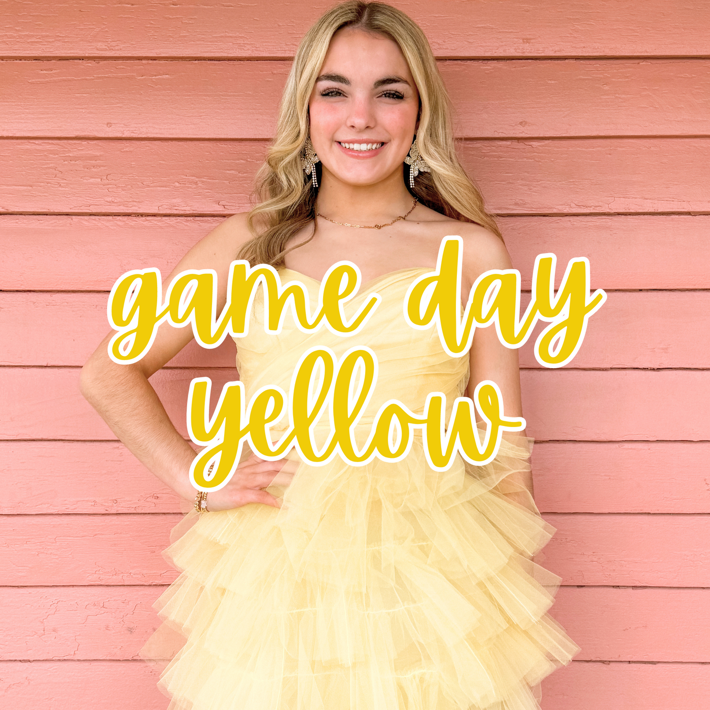 A smiling blonde woman poses against a pink wooden wall, wearing a strapless yellow tulle dress with layered ruffles. "Game Day Yellow" is overlaid in bold yellow script, showcasing a stylish and vibrant outfit perfect for sporting events and team spirit.
