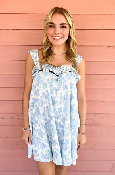 A smiling blonde woman poses against a pink wooden wall, wearing a light blue floral mini dress with ruffle straps and delicate bow details. This feminine and breezy look is perfect for spring and summer occasions.