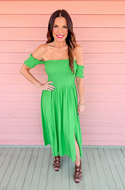 Woman wearing a bright green off-the-shoulder smocked midi dress with short sleeves and side slits. Perfect for spring events, tailgates, or game day. Shop the High Noon Smocked Dress at Style House Frisco for flirty, comfortable, and feminine fashion.